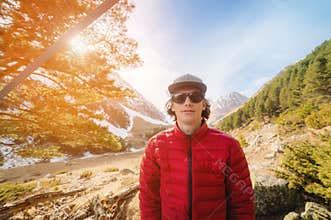 Wide angle Portrait of a long-haired young man in a red down jacket and a gray cap in sunglasses looks into the camera