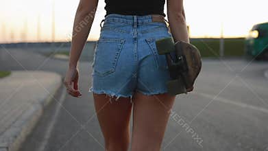 Skateboarder woman carries skateboard at sunset
