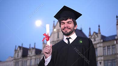 Guy in graduation outfit smiling and showing diploma to camera.