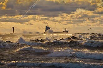 Vessel leaving a harbor and passing lighthouse and pier, breaking through stormy waves, lit by the yellow sunset light