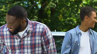 Smiling black dad throwing car keys to teenage son, both getting in vehicle