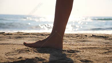 Close up of female feet walking on golden sand at the beach with ocean waves at background. Legs of young woman stepping