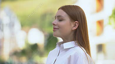 Inspired red-haired teenage girl inhaling pleasant aroma on city street, perfume