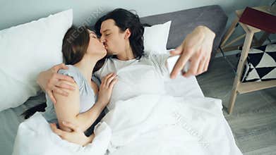 Husband and wife taking selfie with smartphone camera kissing hugging in bed