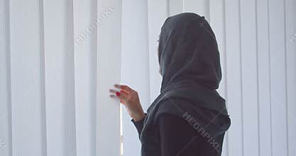 Closeup portrait of young attractive muslim woman in hijab looking through the window with tulle indoors in apartment