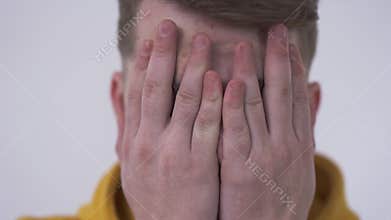 Close-up of a shy young man looking at the camera from behind fingers covering face with his hands. Concept of hiding