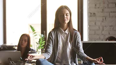 Serene woman sitting at desk meditating while colleagues working