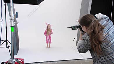 Unrecognizable person with camera taking photos of excited girl in bathrobe jumping on white background in studio