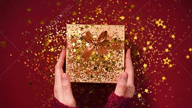 Woman hands opening gift box. Sparkling gold stars, glitter confetti over present. Unpacking gift on red background