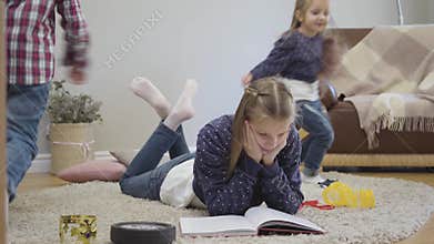 Portrait of cute Caucasian schoolgirl lying on soft carpet and reading as her younger siblings running around. Little