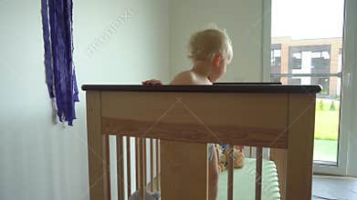 Active baby boy in diapers throwing pillow and toys of his bed. Child clap hands