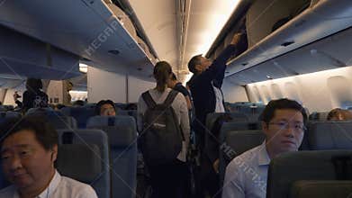 Passengers walk down the aisle, put things up, sit in chairs inside the plane