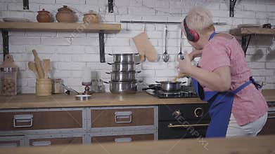 Energetic dance of mature caucasian woman in headphones cooking at the kitchen. Funny senior lady pouring pasta into