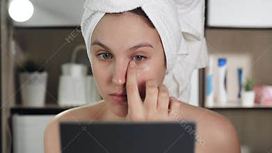 Eye cream. Young attractive woman applies cream to skin under her left eye, girl is in bathroom and looks in mirror