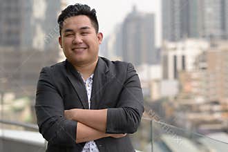 Happy young overweight Asian businessman smiling with arms crossed