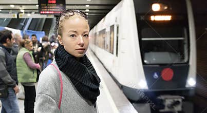 Young woman in winter coat going to work, waiting on the platform of a railway station for train to arrive. Public