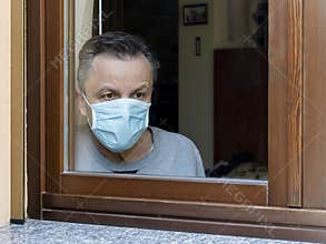 Italian man with protective mask, forced to stay at home due to coronavirus covid-19, disconsolate looks out the window