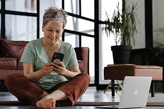 A middle aged lady rests after training yoga in the large hall. A joyful smiling woman sits on a mat on the floor, uses