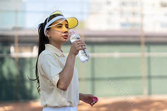 Young woman tennis player on the court drinking water. Recreation after hard exercises. Sportive girl in sun hat drinks