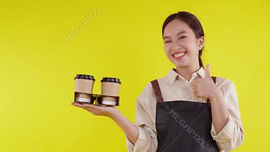 Portrait young asian barista woman wearing apron holding coffee cup and presenting on yellow background.