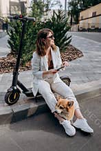 Young businesswoman in white suit sitting on electric scooter and using phone during walking with Welsh Corgi Pembroke dog in city