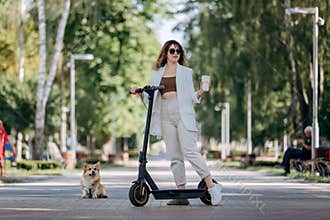 Beautiful young woman in white suit and sunglasses standing with her electric scooter and Corgi Dog in city parkland and drinking