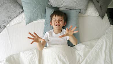 Joyful little boy in bed, opening his blanket and smiling at the camera, showcasing the child's happiness and sense of fun.