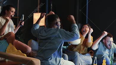 Group of happy young multiethnic friends watchinfg fotball game on TV, eating popcorn and emotionally celebrating win