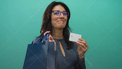 Middle aged hispanic woman holds shopping bag and credit card in studio with turquoise background; confidence
