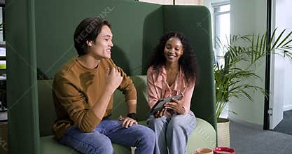 Diverse coworkers entering green booth at office reviewing work on tablet and sipping coffee mug