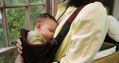 On opening mother is patting sleeping infant and adjusting carrier straps by window for soothing