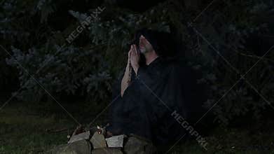 A hooded ascetic monk prays by a fire deep in the forest.