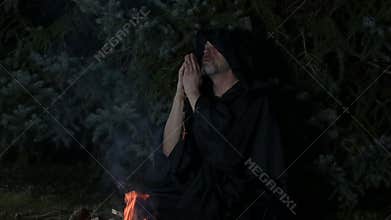 A hooded ascetic monk prays by a fire deep in the forest.
