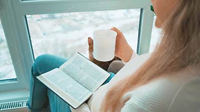 woman with bengal cat reading novel book sitting near window on armchair. Cozy home white room, apartment urban winter