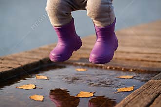 Child in purple boots jumping over autumn puddle on wooden path with scattered yellow leaves, AI Generative