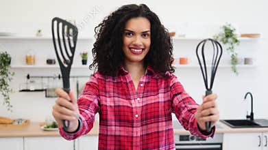 Smiling woman in bright kitchen holding cooking utensils wearing checkered shirt