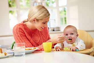 Mother Feeding Baby Sitting In High Chair At Mealtime