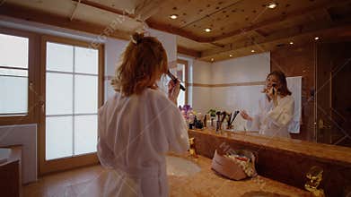 Blonde in bathroom blushing her cheeks dressed in bathrobe.