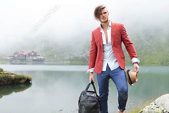 Fashion man holding his hat and travel bag near lake