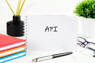 On a clean desk, a spiral-bound notebook displays the handwritten text API. Surrounding it are colorful books, a pen