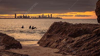 Surfers wait for wave with a sunset under clouds with a city in the distance.
