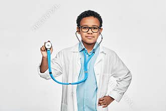 Portrait of African American male kid in a doctor coat with phonendoscope, standing on light grey background