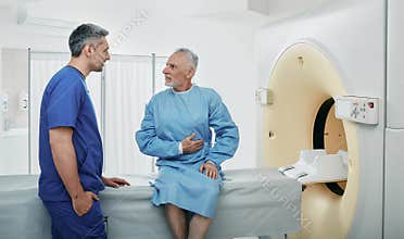 Radiologic technologist listening to senior patient complaints sitting on CT scanner bed. Computed Tomography with contrast