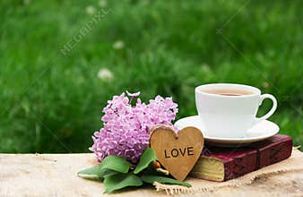 A cup of hot tea, a book and lilacs against a background of green grass. Romantic concept. Wooden card with a heart.