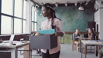 Young african woman getting fired from work. Female walks through the office, carrying box with personal belongings.