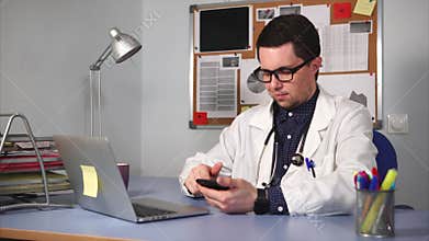Male medic in white coat with stethoscope sitting at desk and using smart watch.