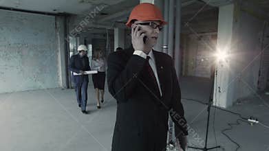 Man in suit, helmet call inside construction site. At background team of young builders man, african