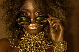 Close-up of magic golden african american female model in massive sunglasses with bright glitter makeup, glossy