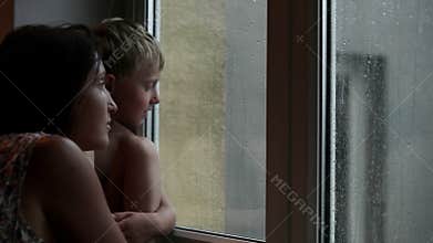 Mother and son awaiting someone to come looking out the window during the rain