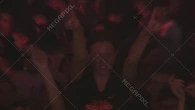 Crowd of people dance in nightclub. Girl sit on shoulders and show fingers up. DJ at turntable